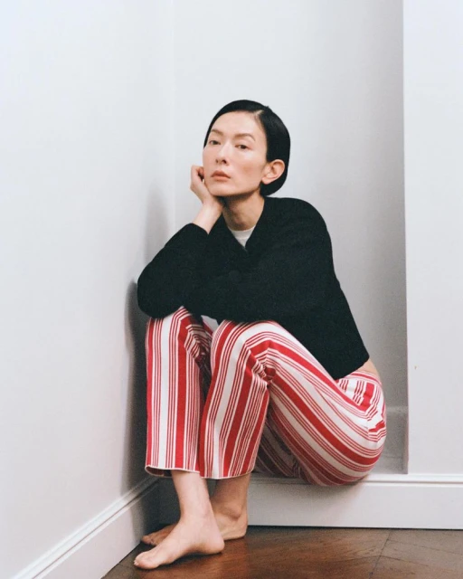 Woman seated in corner wearing striped pants and a sweater, thoughtfully gazing upward, creating a portrait in a simple indoor setting.