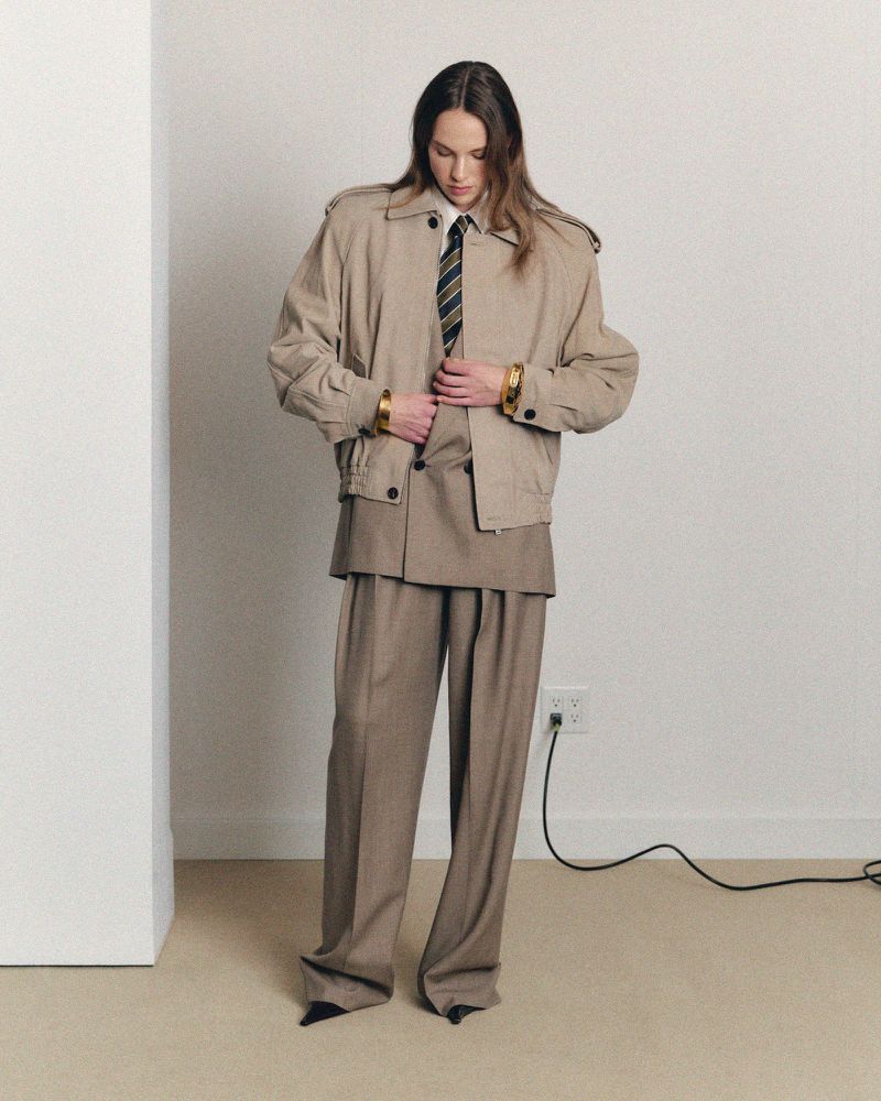 Woman poses in an oversized beige suit with jacket and tie in a minimalist studio setting, presenting a unique fashion statement.