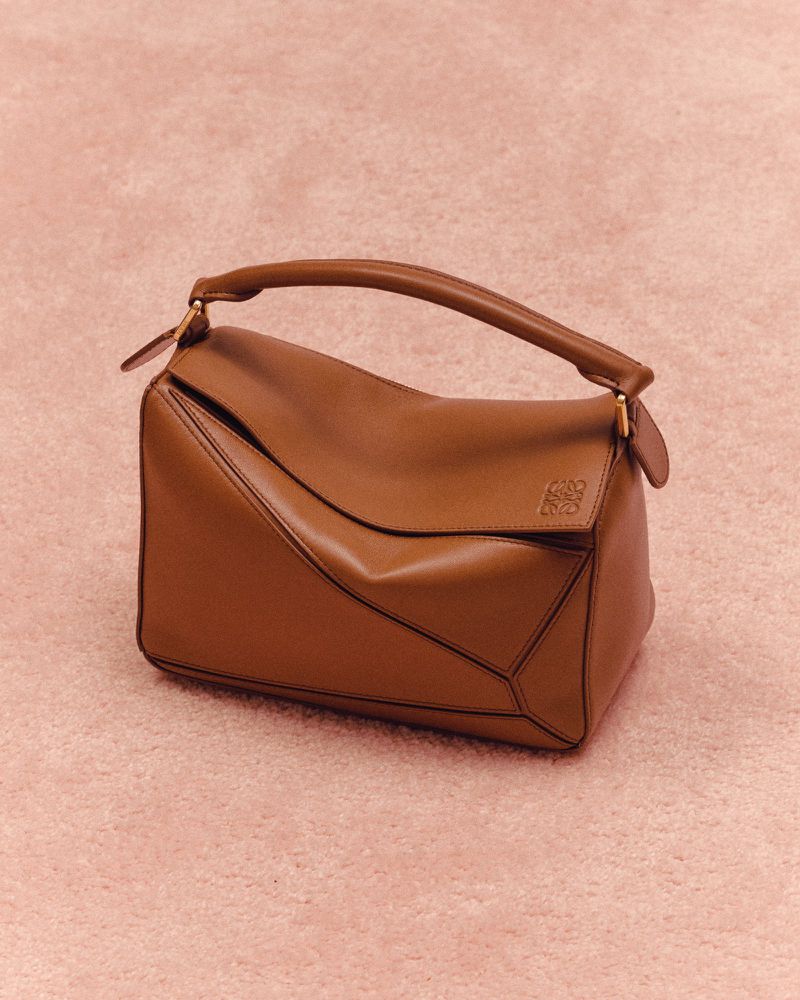Studio shot of a brown Loewe Puzzle bag with a top handle against a light pink textured background, showcasing modern fashion.