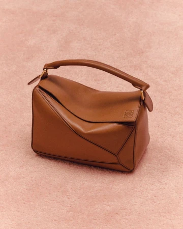 Studio shot of a brown Loewe Puzzle bag with a top handle against a light pink textured background, showcasing modern fashion.