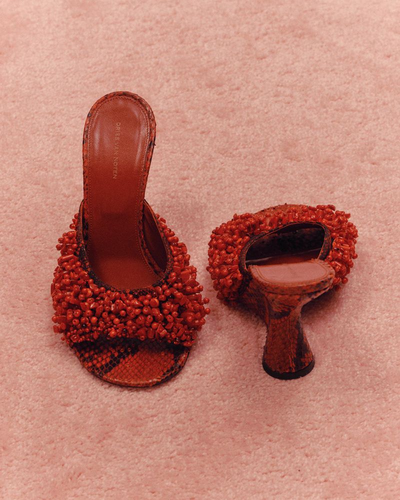 Elevated view of two red, beaded high-heel mule shoes on a light-red carpet, showcasing fashion footwear and textured details.