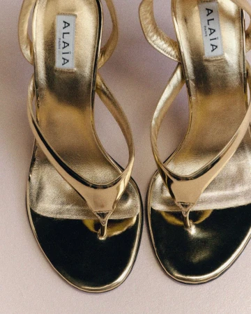 Close-up of gold leather sandals with thin straps, featuring the Alaïa brand logo on the inner sole, set against a neutral background.