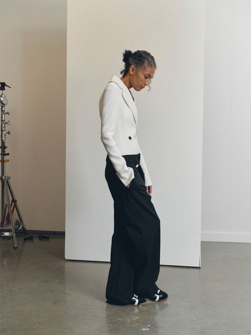 Woman poses in a photo studio wearing a white blazer, black wide-legged trousers, and black and white sneakers against a neutral backdrop.