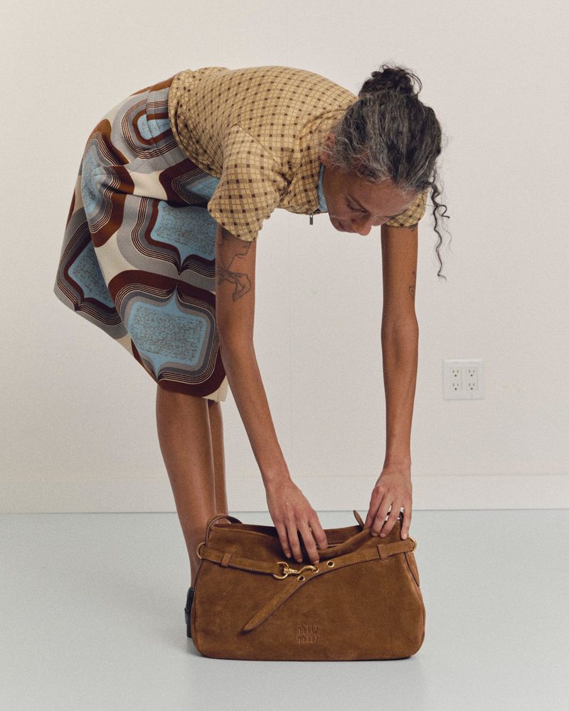 Adult woman with gray hair leans over brown suede bag, dressed in a patterned skirt and short-sleeved shirt against a white background.