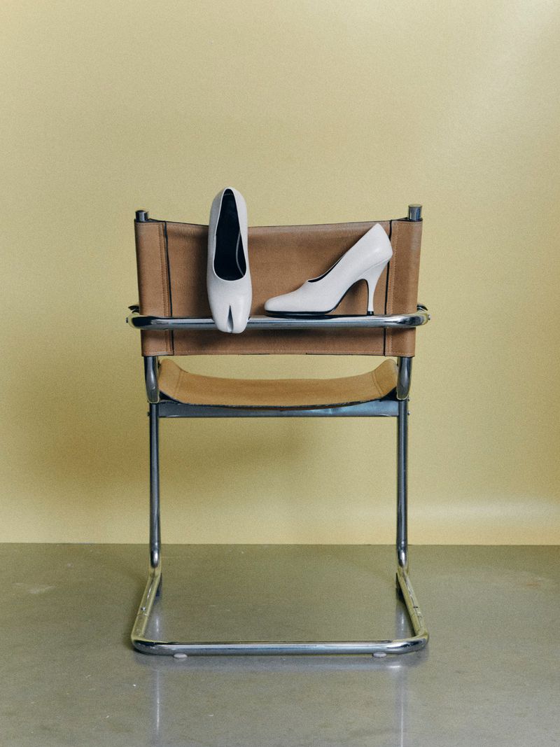 Two white high-heel shoes, one standing upright and the other lying on its side, are placed on a metal and tan leather chair.