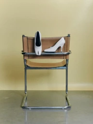 Pair of white high heels resting on a tan leather and chrome tubular chair against a solid pale yellow backdrop