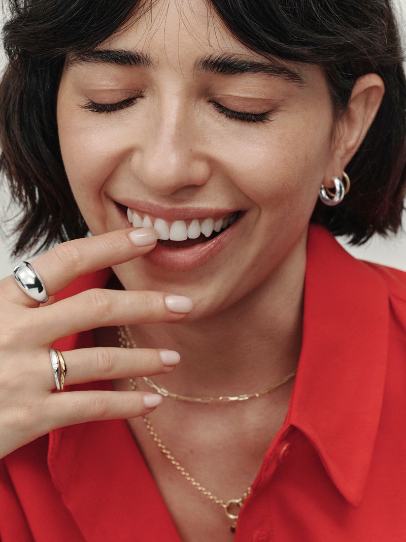 Close-up portrait of a smiling woman wearing layered gold necklaces, silver hoop earrings, and rings for Mejuri Art Of The Stack.