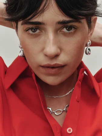 Close-up of a brunette woman with a serious expression, wearing silver jewelry and a bright red collared shirt against a neutral background.