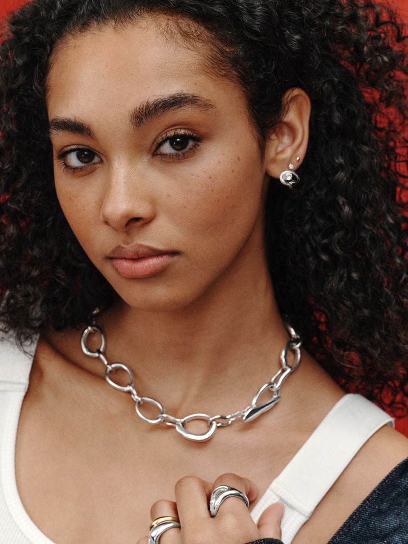 Close-up of an adult female with curly brown hair modeling a silver chain necklace and several silver rings against a red background.