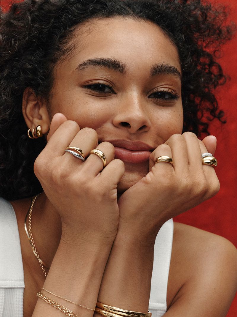 Close-up of a young woman with curly hair, smiling, wearing multiple gold rings on her fingers and gold jewelry, against a red backdrop.