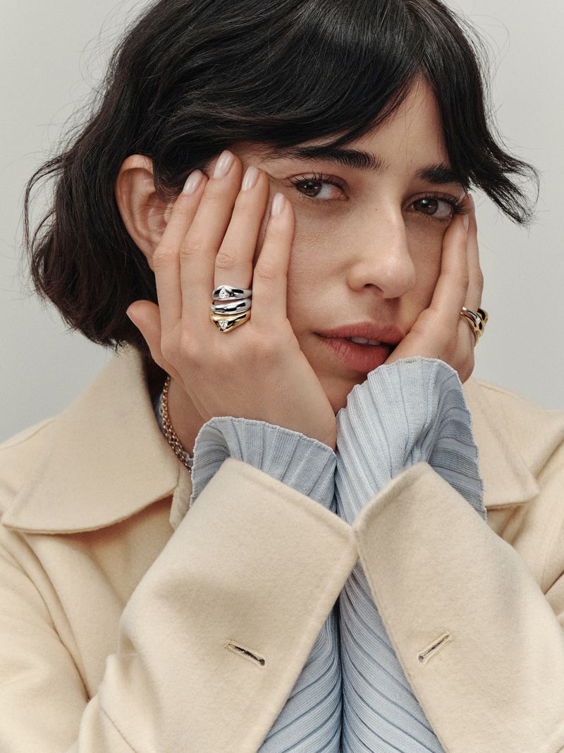 Close-up of a brunette woman with fair skin wearing gold and silver rings, a light blue shirt, and a beige coat, looking at the camera.