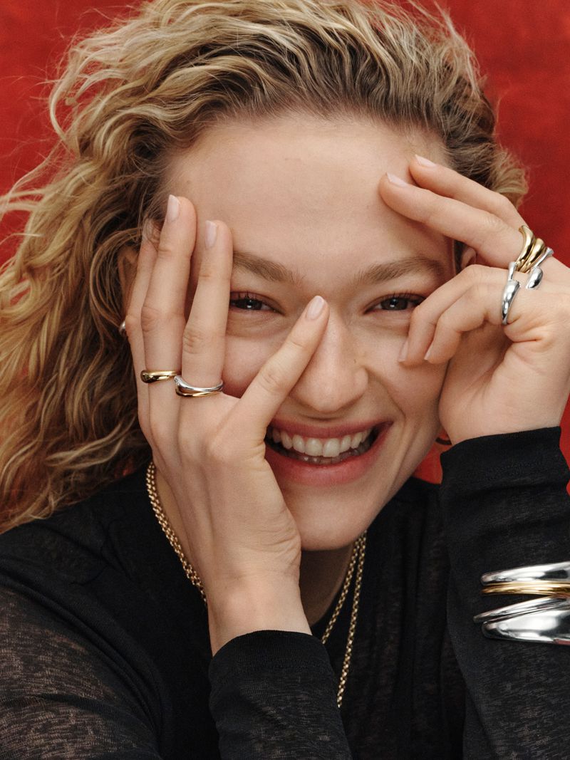 Close-up portrait of a smiling woman wearing multiple rings and bracelets against a textured red backdrop.