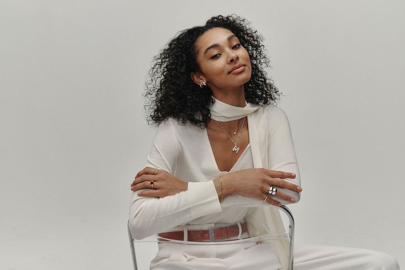 Attractive young woman with curly hair poses in a light studio wearing jewelry, a white top, and pants while sitting on a clear chair.