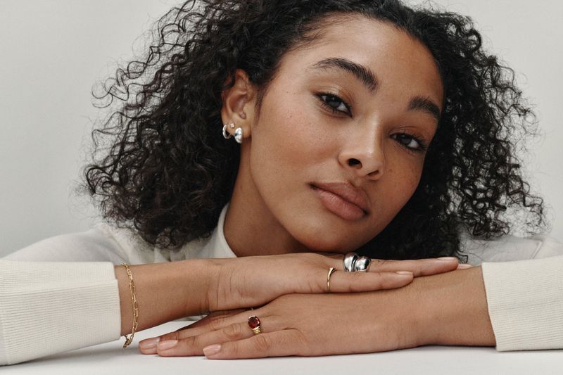 Close-up studio shot of a curly-haired woman with rings and a bracelet, showcasing jewelry against a neutral background.
