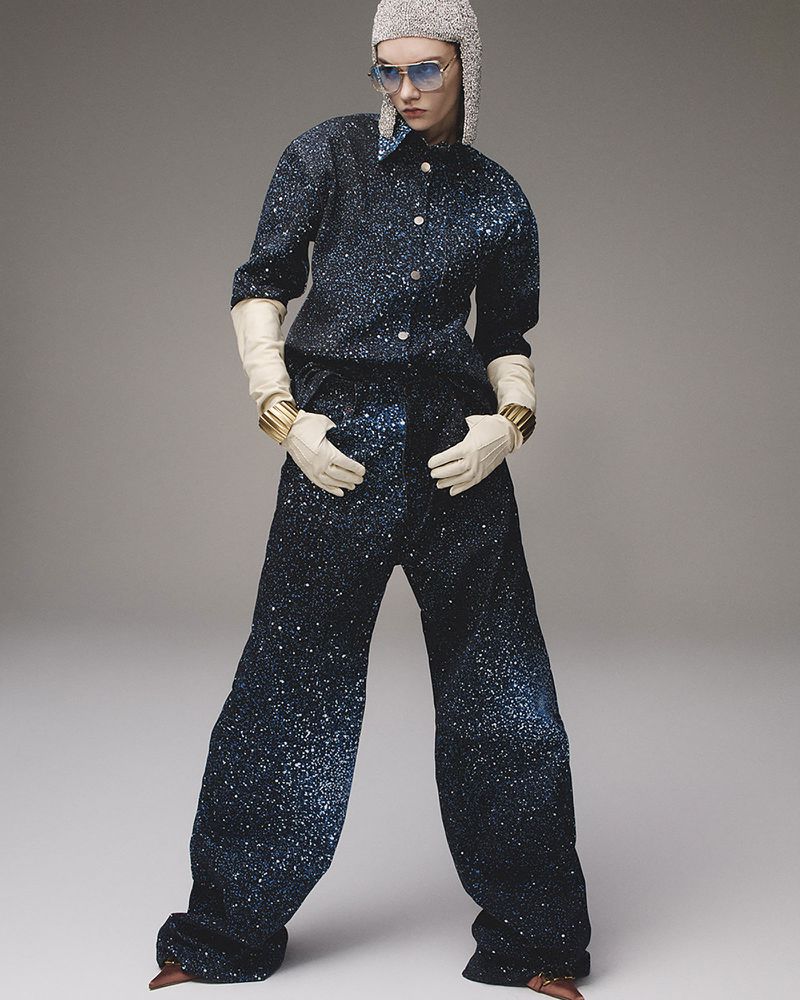 Fashion editorial shot of a female model wearing a sparkly blue outfit, white gloves, silver hat, and glasses in a studio setting.