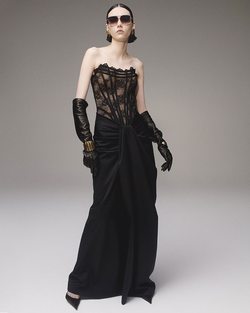 Elegant adult female fashion model poses in a black lace corset top and long skirt, wearing opera gloves, sunglasses, and gold jewelry.