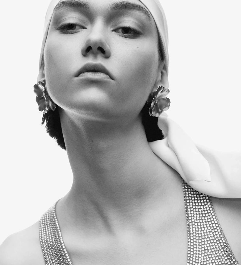 Black and white portrait of a woman with floral earrings and a headscarf, set against a stark white background. Fashionable and elegant.