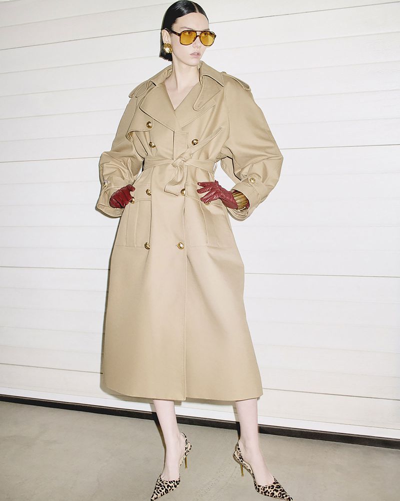 Model wearing a belted beige trench coat, yellow-tinted sunglasses, and leopard print heels against a white paneled wall.