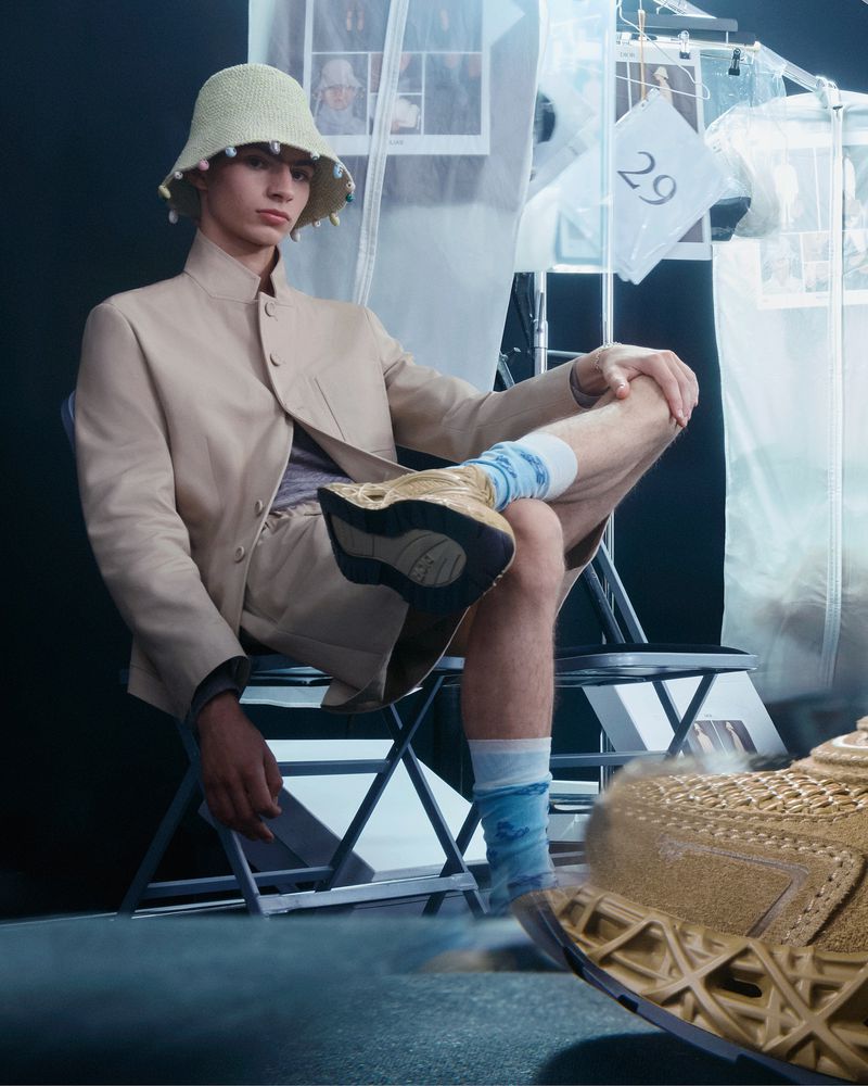 Young adult man in casual fashion seated on a folding chair, wearing a bucket hat and socks, paired with beige sport shoes.
