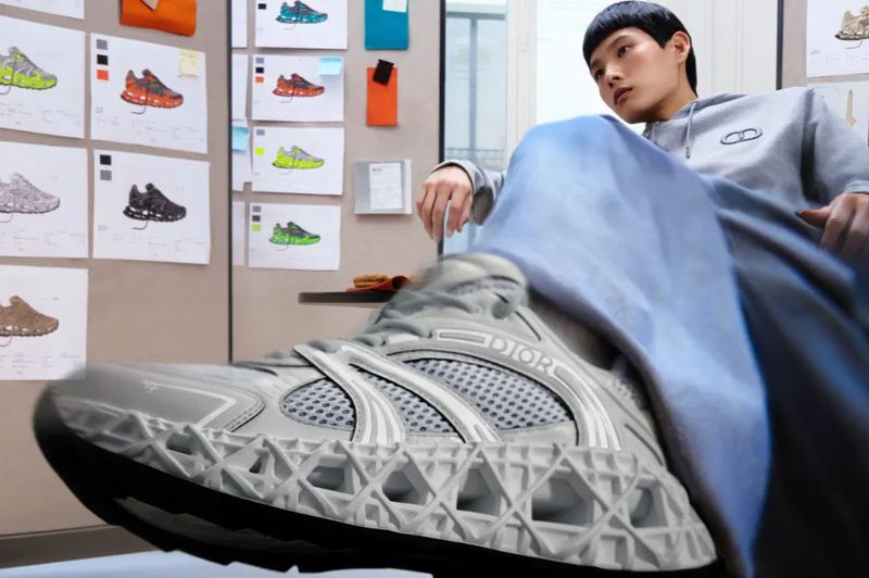 A light-skinned male model in a grey hoodie sits with a Dior sneaker prominently displayed; shoe sketches line the background wall.