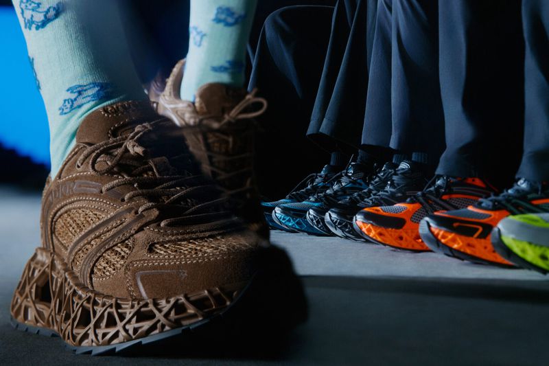Detailed shot of a brown sneaker with a complex sole design, alongside a lineup of colorful sneakers and people wearing matching dark pants.