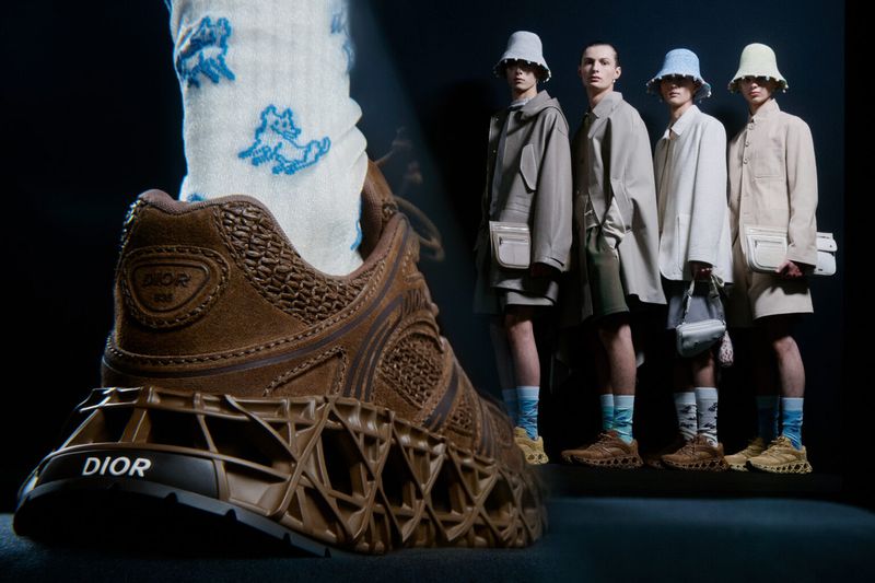 Brown Dior sneaker on display, featuring a sock with blue designs, with four models in coordinated attire standing in background.