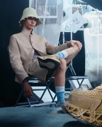 Model seated on a folding chair wearing a beige jacket and bucket hat with hanging beads, photographed by Matthew Tammaro for Dior Men.