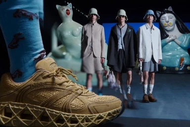 Close-up of a tan suede sneaker in foreground with three male models standing in the background beside large ceramic cat sculptures.