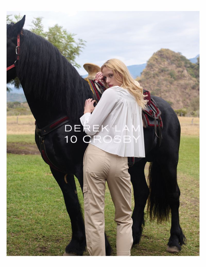 Adult female poses next to black horse in outdoor fashion shot with field and mountain background; shirt, pants, saddle shown.