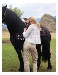 Blonde model wearing a white top and tan trousers standing beside a black horse in an outdoor field.