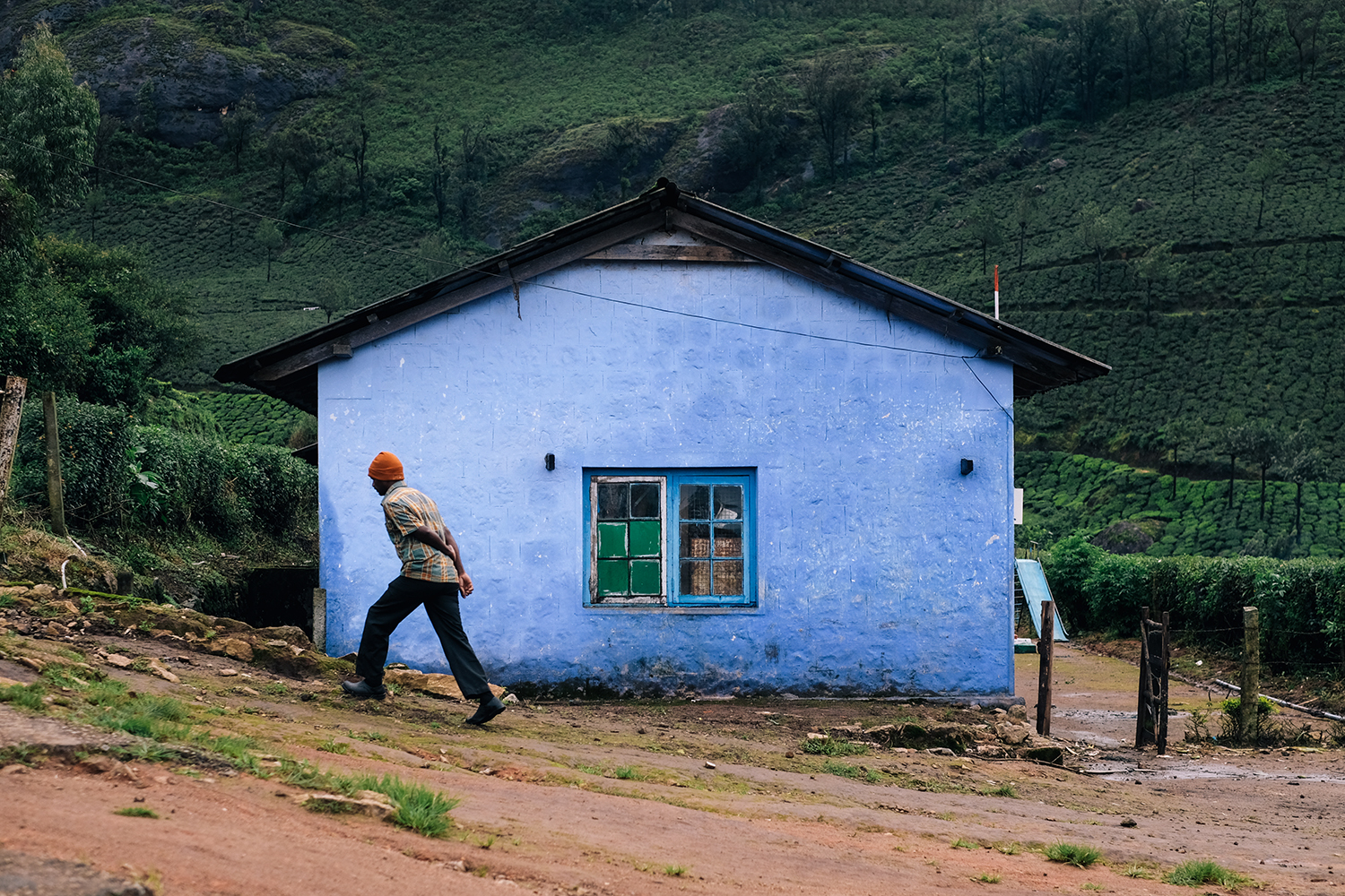 A person walks on a path leading to a light-blue house, set against a lush green hillside, presenting a serene rural scene.