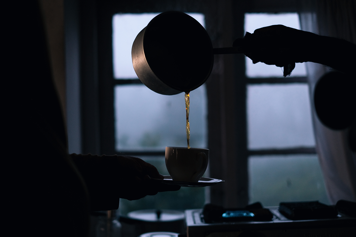 Silhouette image of a person pouring a liquid from a metal pot into a cup, with window and foggy landscape in the background.