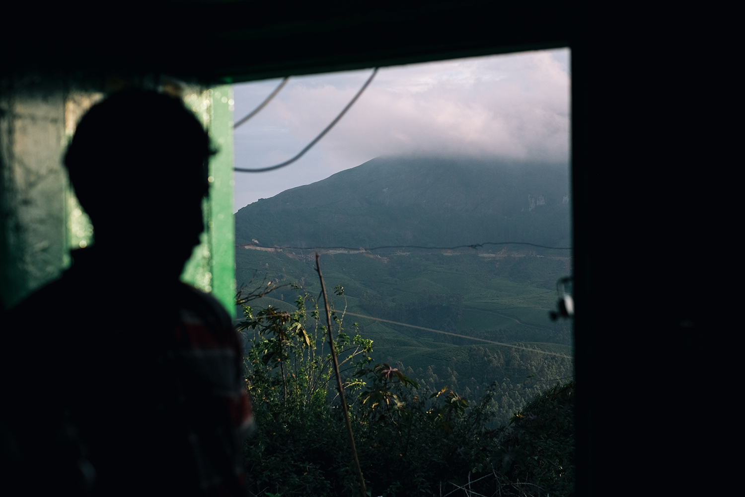 A silhouette of a person looking at a mountain range with green slopes, partially covered in clouds, viewed through a doorway.