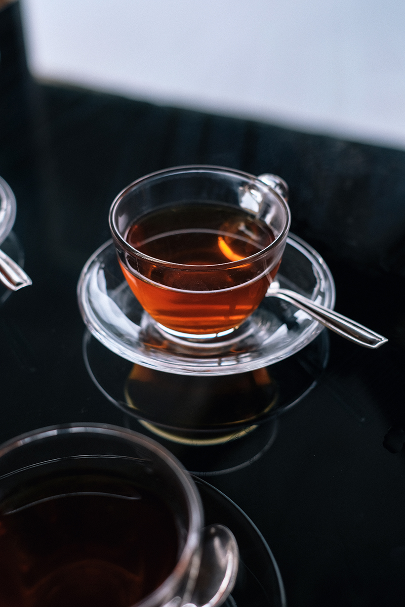 Close-up of two clear glass cups filled with amber liquid on saucers, one cup in focus, set on a glossy black surface.