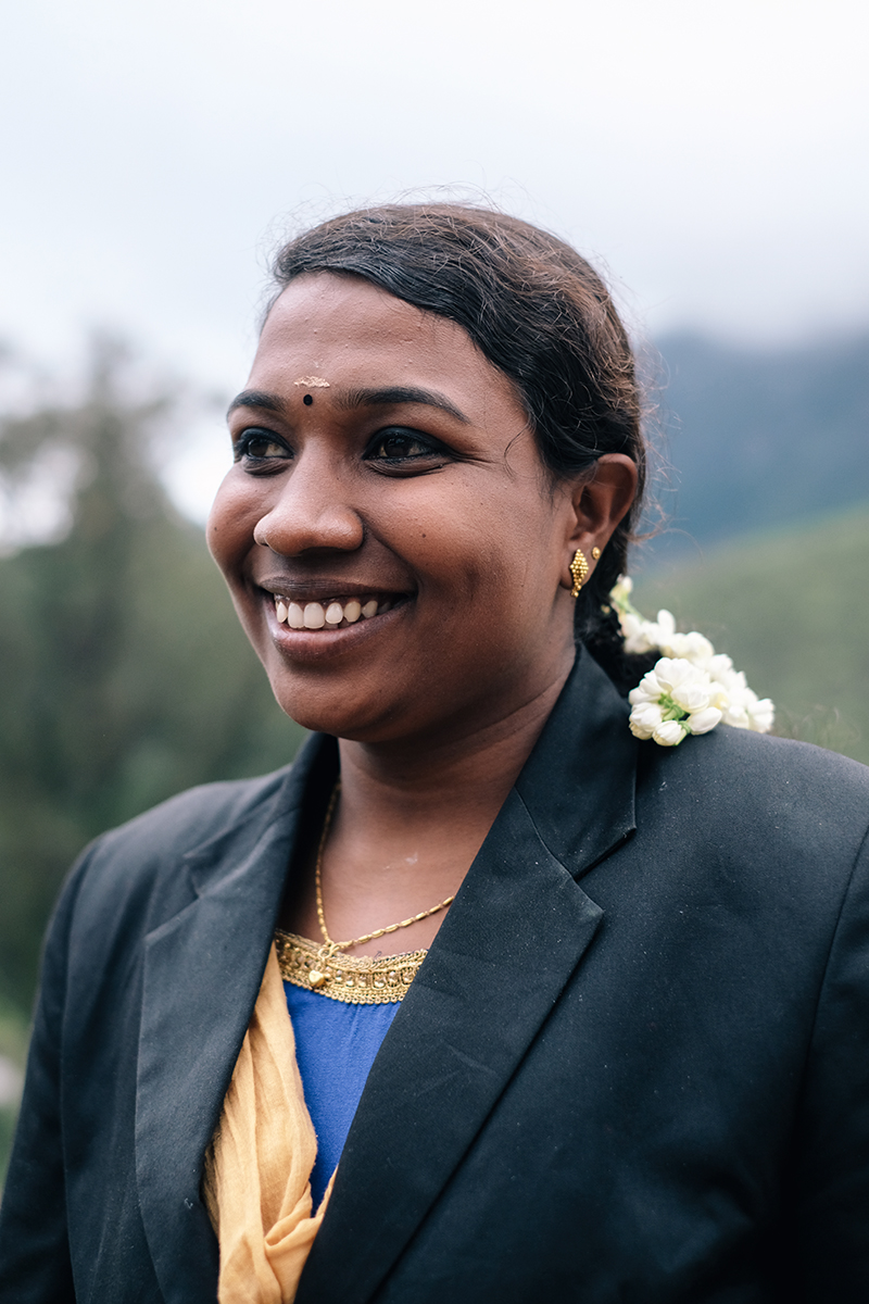 Smiling Indian woman in black blazer, wearing flowers in her hair, against a soft-focus green background of trees and cloudy sky.