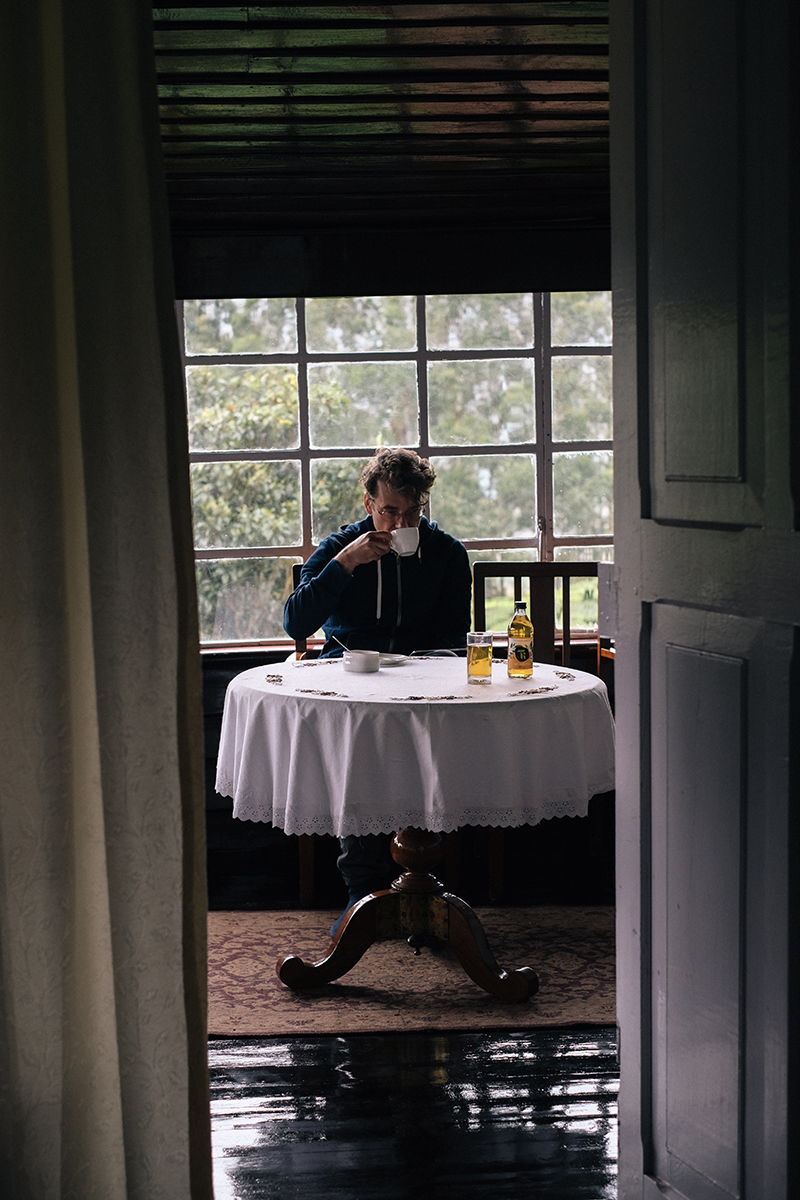 Man drinks from a white cup while sitting at a table with a white tablecloth near a window, viewed through a doorway from a dark room.