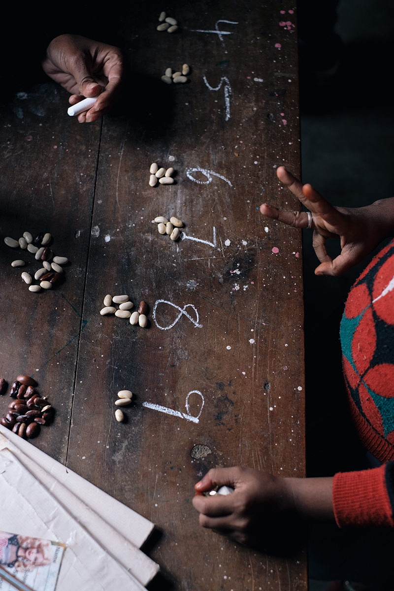 Wooden table surface featuring the numbers four through nine written in chalk with groupings of beans beside each number, and hands present.