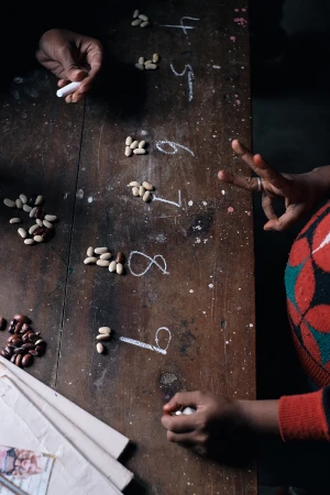Wooden table surface featuring the numbers four through nine written in chalk with groupings of beans beside each number, and hands present.