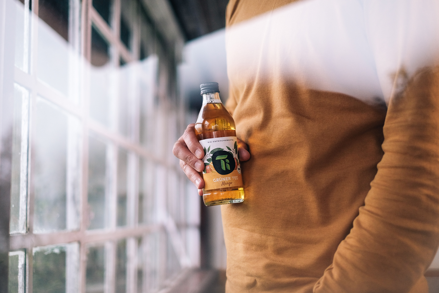 A person holding a glass bottle of Ti green tea next to a large multi-pane window with soft natural light.