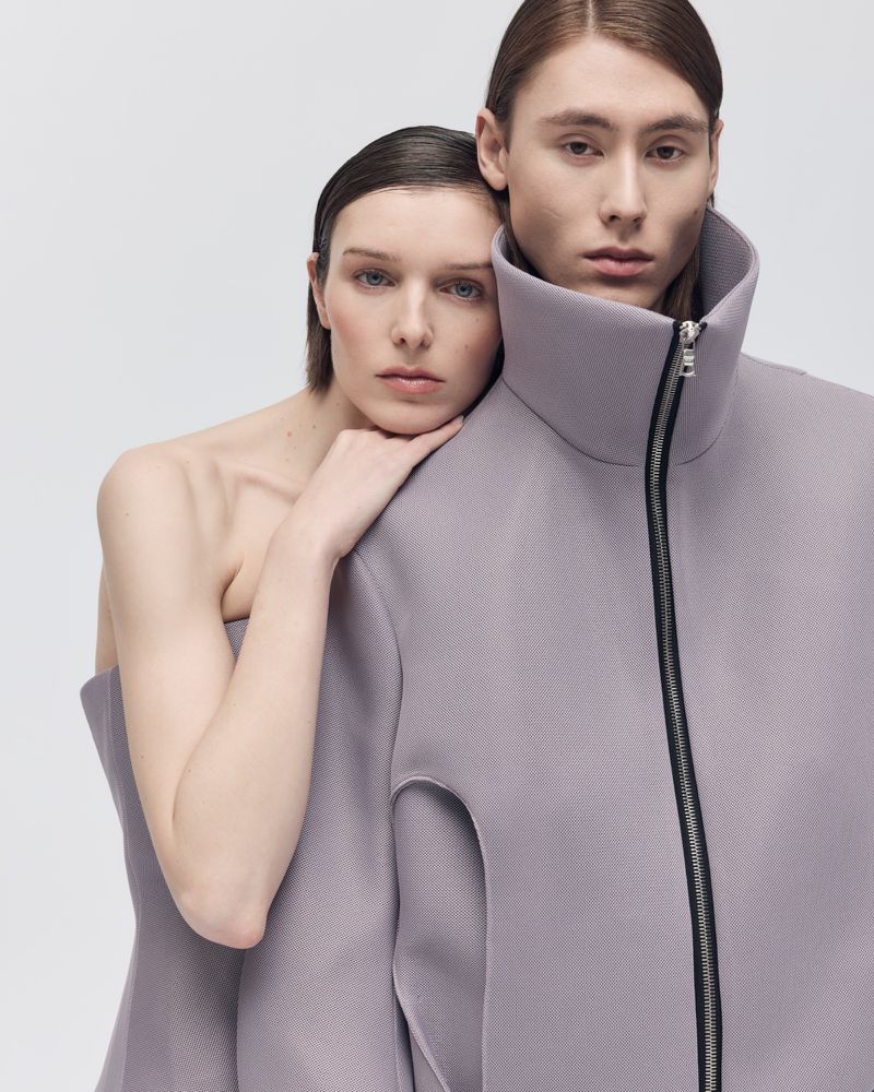 Two fashion models posing in a studio. The female model is leaning on the male model. They are wearing purple structured clothing.