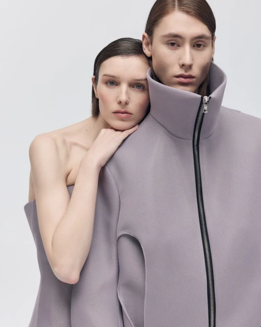 Two fashion models posing in a studio. The female model is leaning on the male model. They are wearing purple structured clothing.