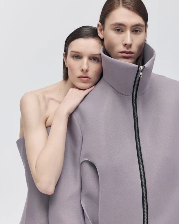 Two fashion models posing in a studio. The female model is leaning on the male model. They are wearing purple structured clothing.