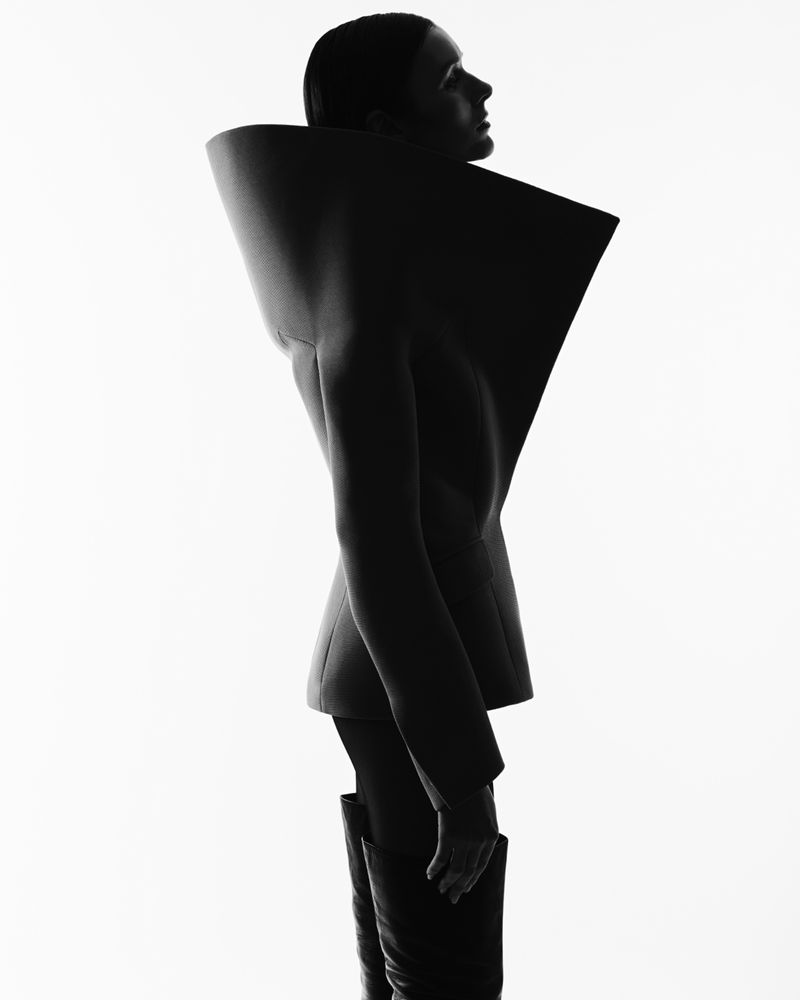 Monochrome studio shot of a slim adult female in dark blazer with large collar and dark boots, standing in profile against a white background.