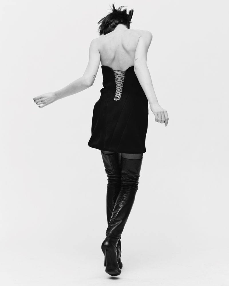 Back view of a woman in a black strapless dress and thigh-high boots against a white background, in a black and white studio shot.