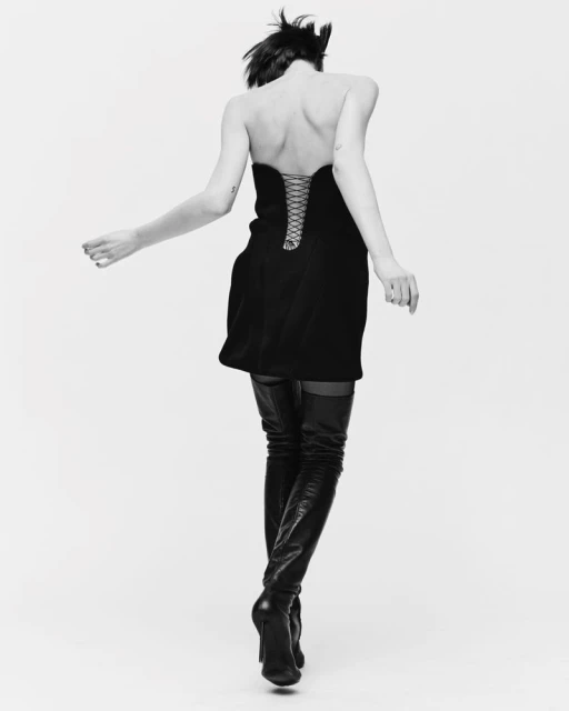 Back view of a woman in a black strapless dress and thigh-high boots against a white background, in a black and white studio shot.