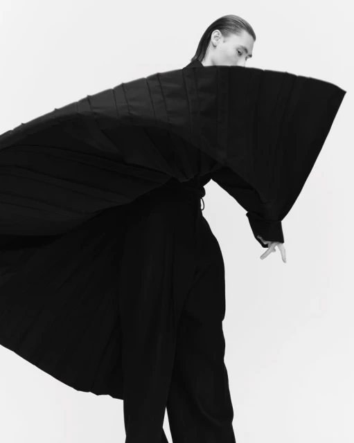 Monochrome studio shot of a man posing in a wide-sleeved black robe and loose pants against a white background. Fashion editorial.