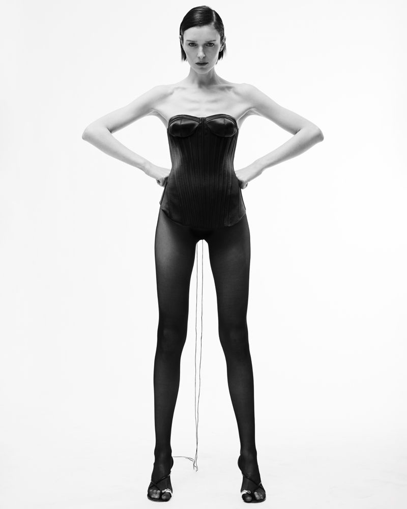 A striking black and white studio shot captures a woman in a dark corset and sheer tights, her hands on her hips, against a seamless white backdrop.