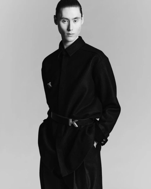 Monochrome portrait of a slender adult male model in dark, draped clothing with a 'K' belt buckle, set against a neutral backdrop.