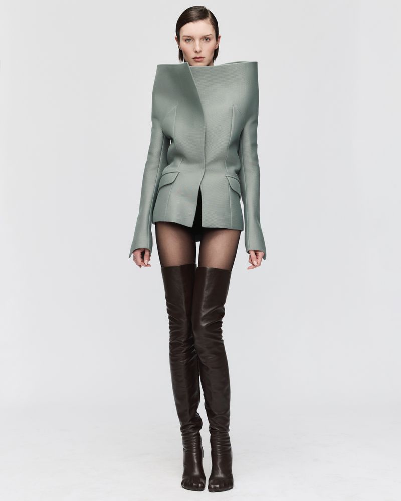 Fashion model in studio wearing an avant-garde blazer and thigh-high boots against a stark white background in a fashion editorial shot.