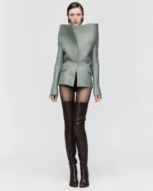 Fashion model in studio wearing an avant-garde blazer and thigh-high boots against a stark white background in a fashion editorial shot.